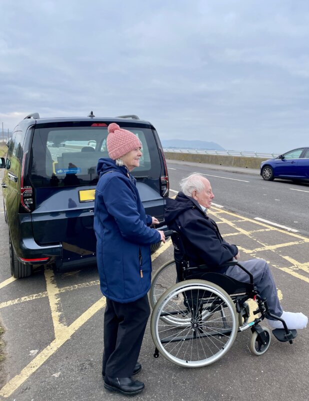 Smiling man in wheelchair being pushed by smiling woman, next to blue car.